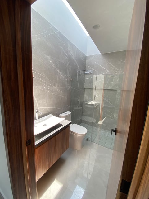 Modern bathroom with gray tile walls, frameless glass shower enclosure, white sink vanity, toilet, and skylight above.