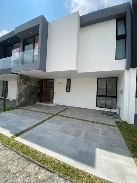 Modern white and gray two-story residential house with glass balcony, paved driveway, and manicured lawn