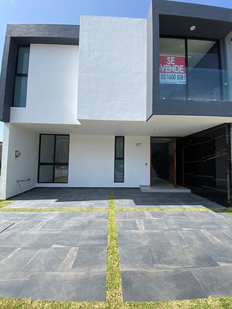 Modern two-story white house with black accents, garage, and for sale sign. Contemporary residential architecture with geometric design.