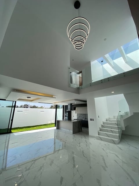 Modern home interior with high ceiling, geometric pendant light, marble flooring, and white staircase visible on right side