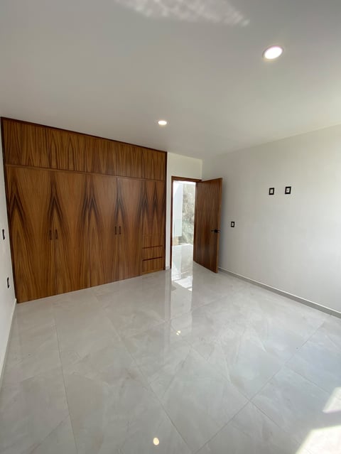 Empty room with white polished floor, wooden wall paneling with doors, white walls, recessed ceiling lights, and doorway with natural light