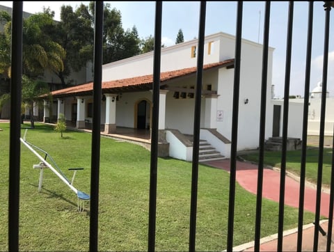 View through black metal fence of white and orange building with green lawn and brick courtyard