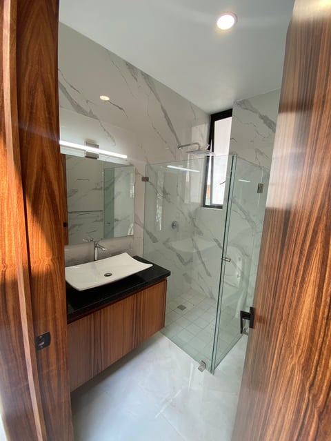Modern bathroom with marble walls, wood vanity with white sink, glass shower enclosure, and recessed lighting.
