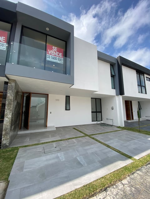 Modern white townhouse with black trim and se vende real estate sign, paved courtyard, and cloudy sky