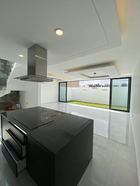Modern kitchen with black island, stainless steel range hood, white walls, and large windows overlooking green landscape.