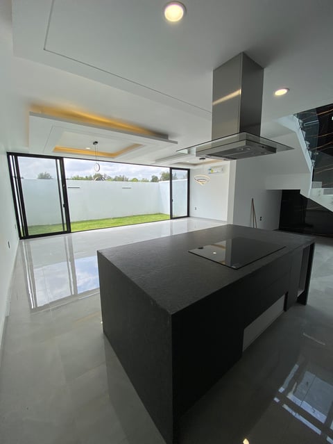 Modern kitchen with black island, stainless steel range hood, white polished flooring, and large windows overlooking green landscape