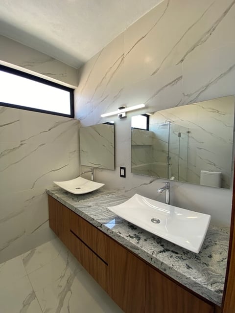 Modern bathroom with marble tiles, dual white vessel sinks, wooden vanity, glass shower enclosure, and skylights